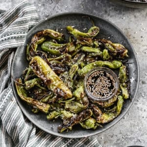 A plate filled with the best Blistered Shishito Peppers, drizzled with soy sauce, ginger, and honey, next to a sesame dipping sauce.
