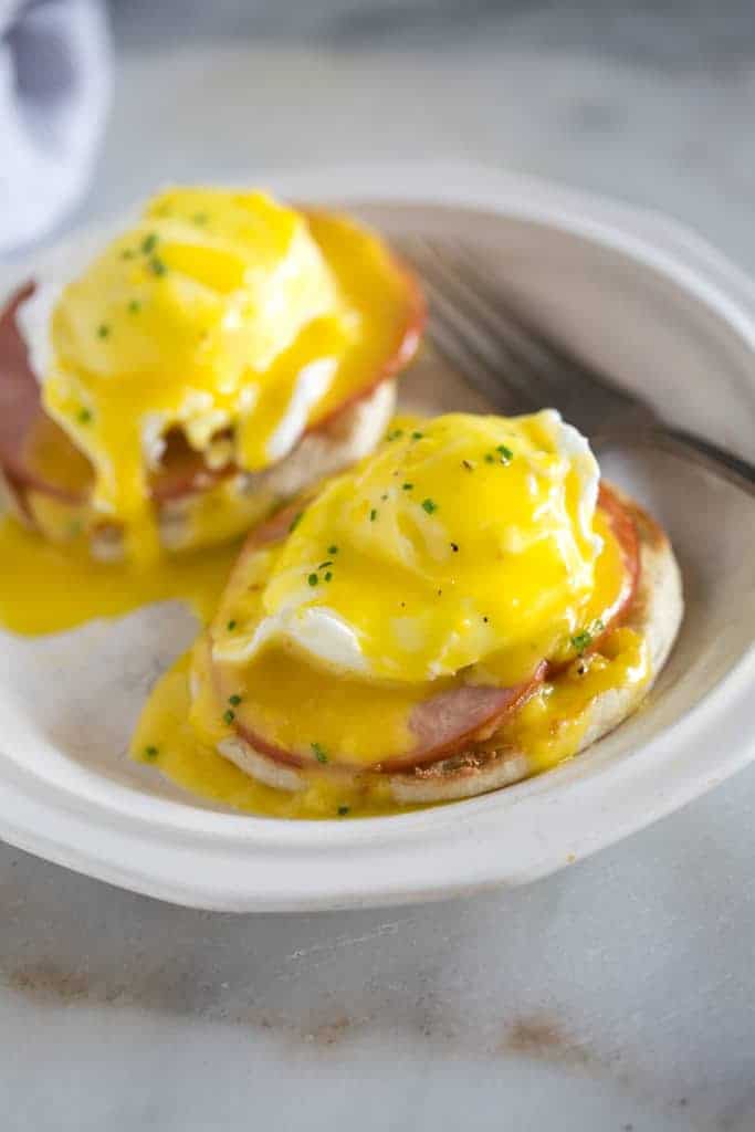 A plate with two eggs benedict which includes english muffin topped with a slice of canadian bacon, poached eggs and homemade hollandaise sauce.