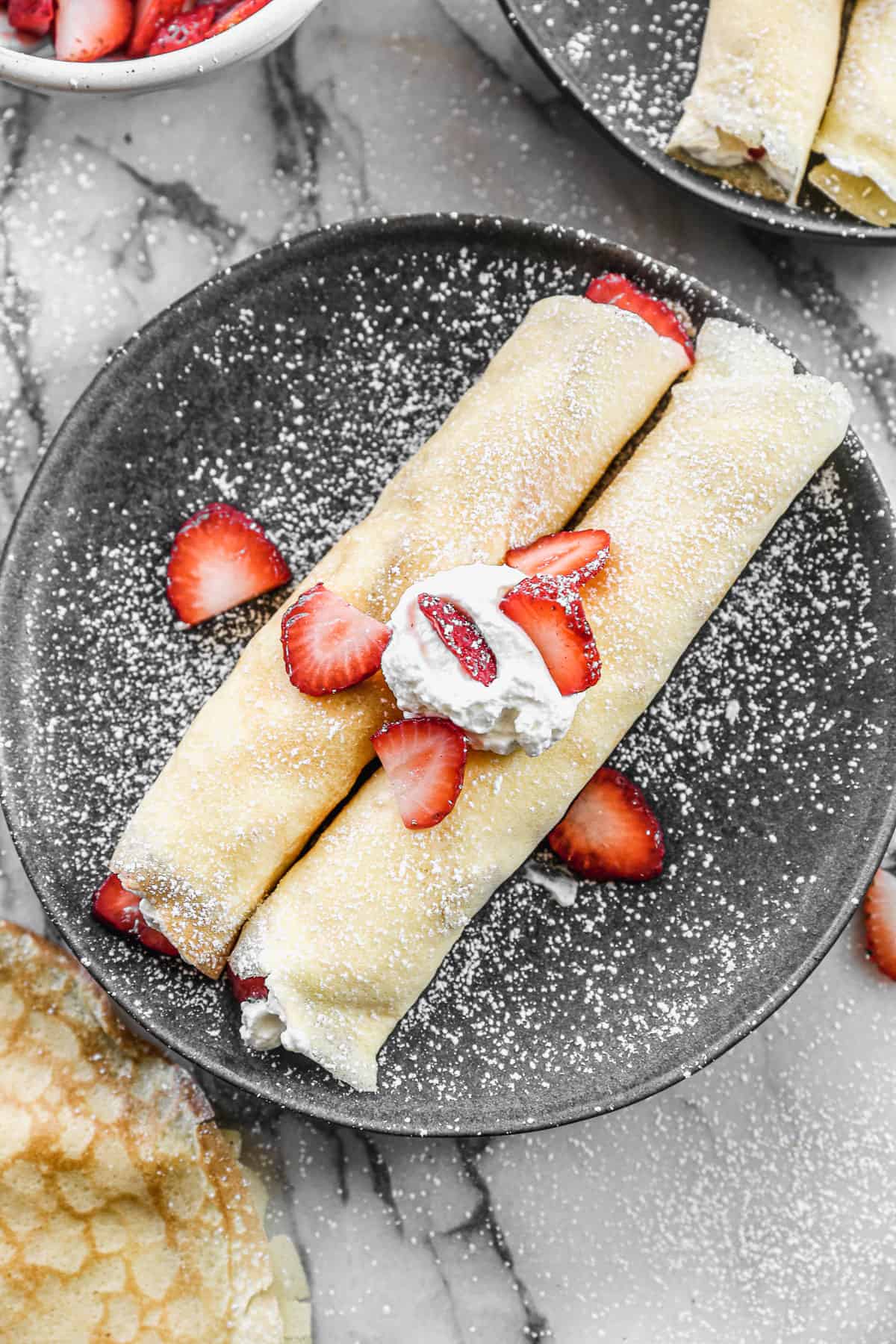 Two of the best Strawberry Crepes rolled up with a spoonful of whipped cream on top and sliced strawberries, all dusted with powdered sugar.
